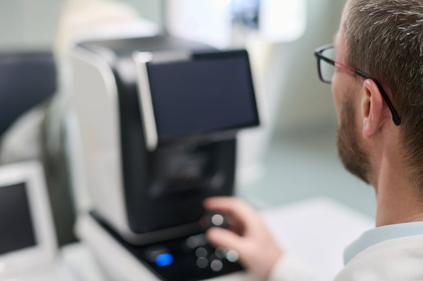 man operating ophthalmic diagnostic device in clinical setting, using control knob while sitting in front of medical equipment during eye examination procedure