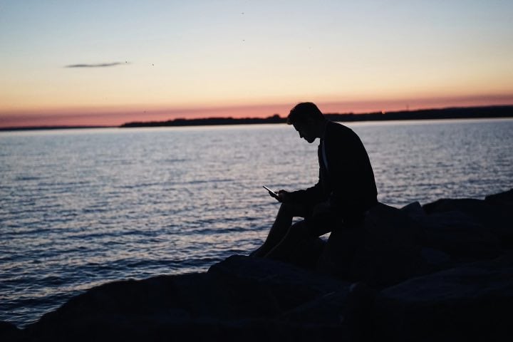 Man looking at his cell phone while sitting next to the ocean